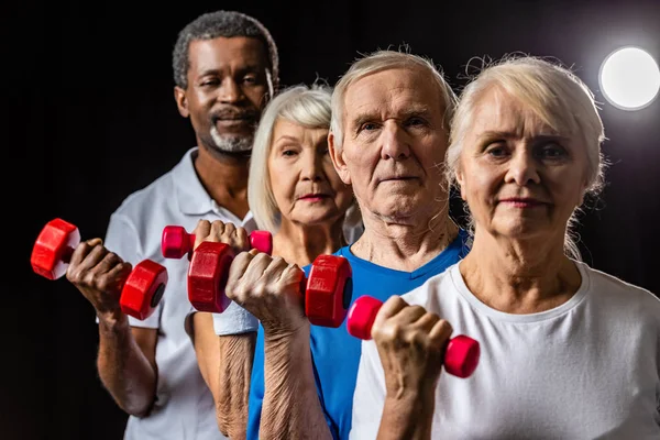 Senior Mutliethnic Sportspeople Synchronous Exercising Dumbbells Black Spotlight — Stock Photo, Image