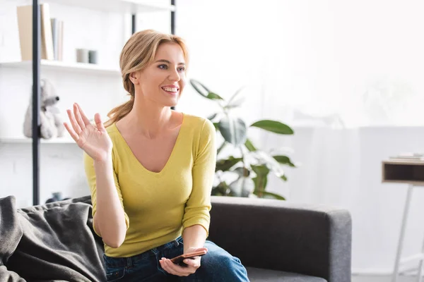 Vackra Leende Kvinna Anläggning Smartphone Och Viftande Hand Medan Sitter — Stockfoto