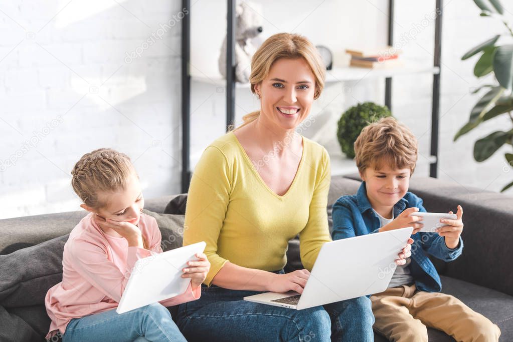 happy mother using laptop and smiling at camera while kids using digital devices at home
