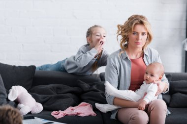 exhausted mother holding infant child and looking at camera while naughty daughter playing behind clipart