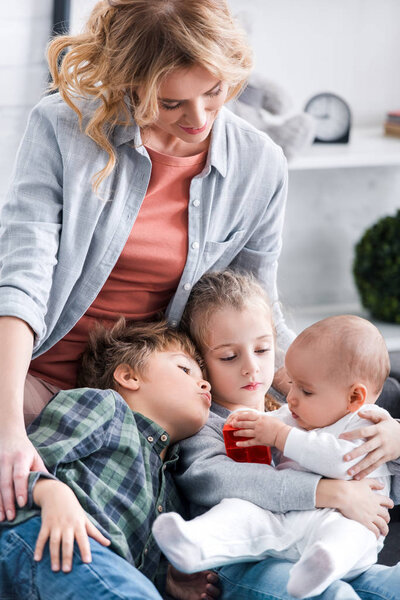 beautiful happy mother looking at three adorable kids playing at home