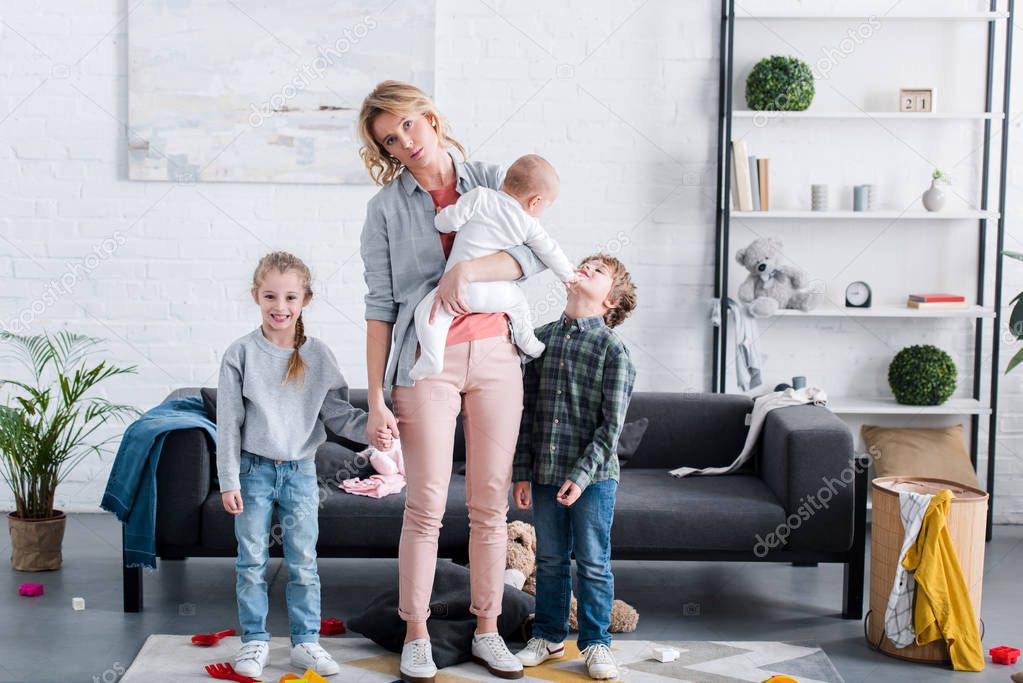 tired mother with three little kids standing in room and looking at camera