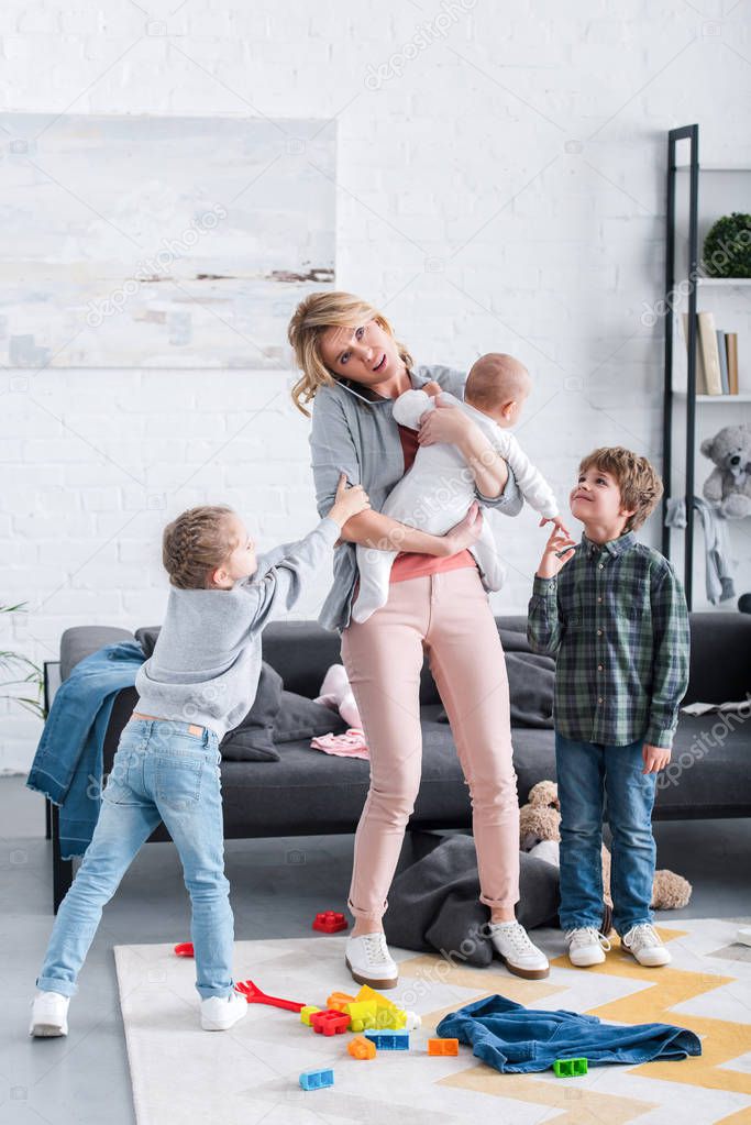 exhausted mother talking by smartphone and holding infant child while siblings playing in room  