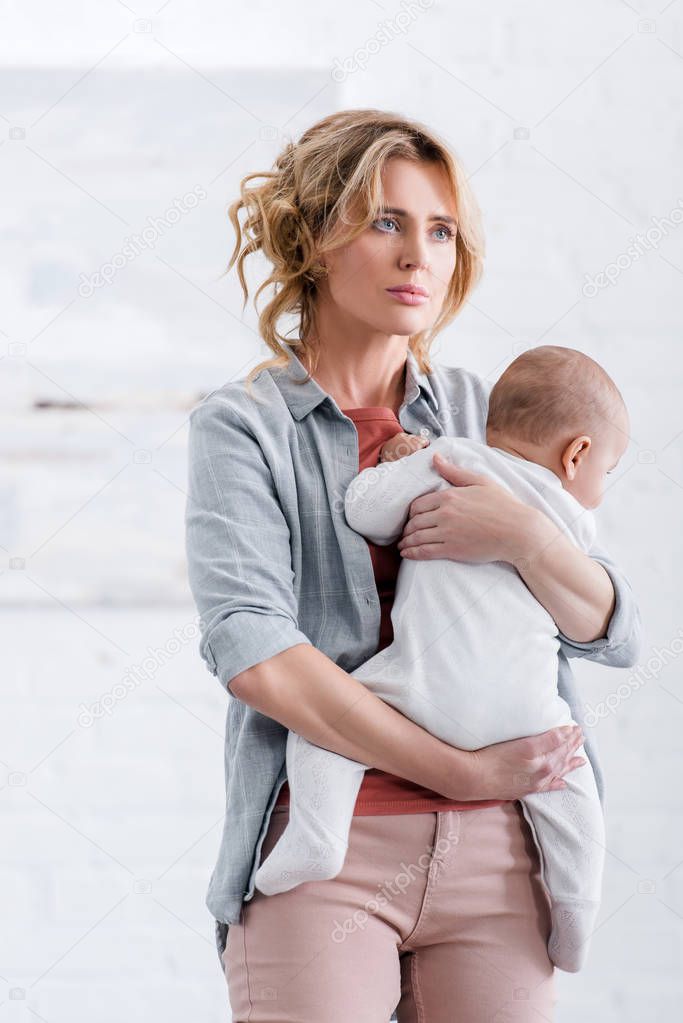 tired mother holding cute infant child and looking away at home
