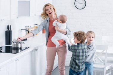 exhausted mother with infant kid cooking and looking up while naughty children playing in kitchen  clipart