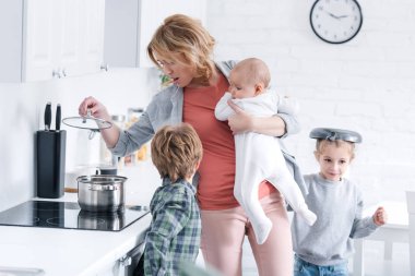 exhausted mother holding infant kid and cooking while naughty children playing in kitchen  clipart