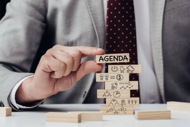 cropped view of man holding brick with word 'agenda' over wooden blocks with icons clipart
