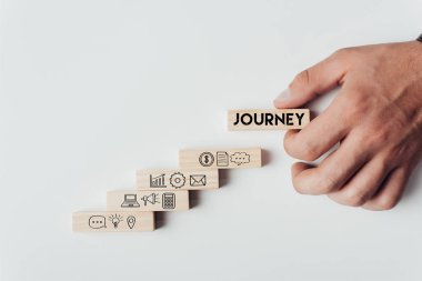 cropped view of man holding wooden block with word 'journey' on top of wooden bricks with icons isolated on white