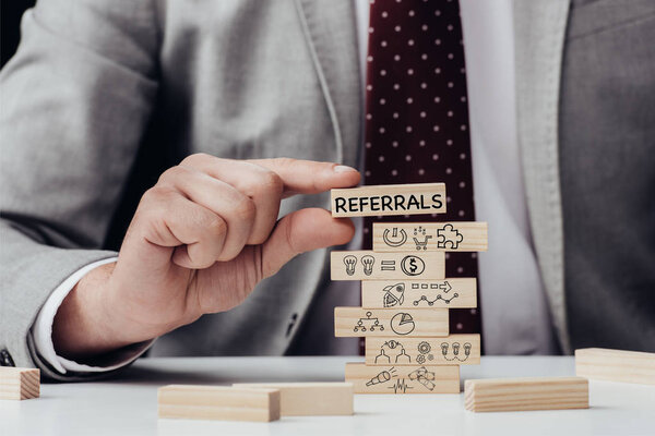 cropped view of man holding brick with word 'referrals' over wooden blocks with icons