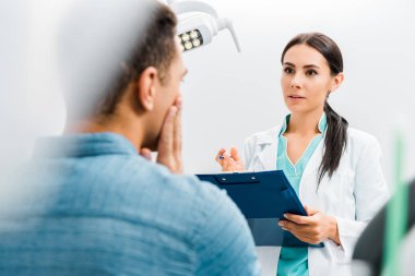 selective focus of female dentist standing with clipboard and talking with african american patient having toothache  clipart