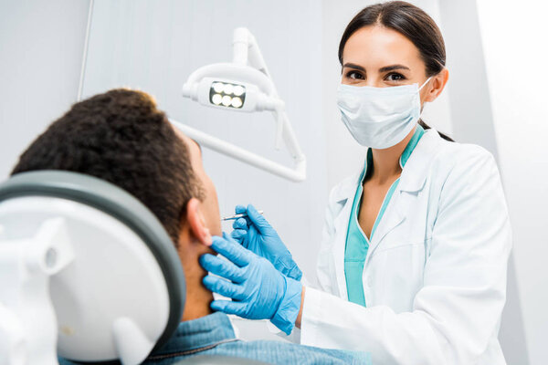 female stomatologist  in latex gloves and mask examining african american patient 