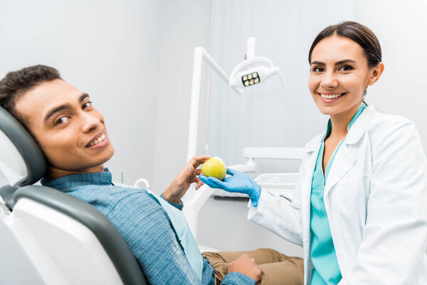 cheerful dentist giving apple to african american patient 