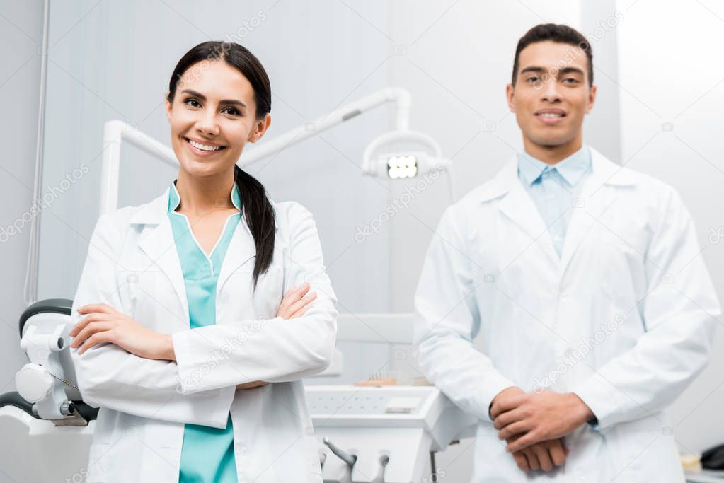 cheerful  female doctor standing near african american coworker in white coat with crossed arms 