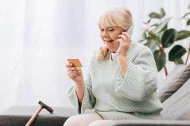 smiling blonde retired woman looking at credit card while talking on smartphone clipart