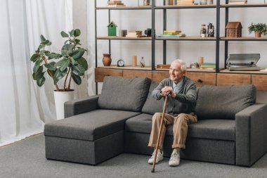 happy senior man smiling and holding walking cane while sitting on sofa  clipart