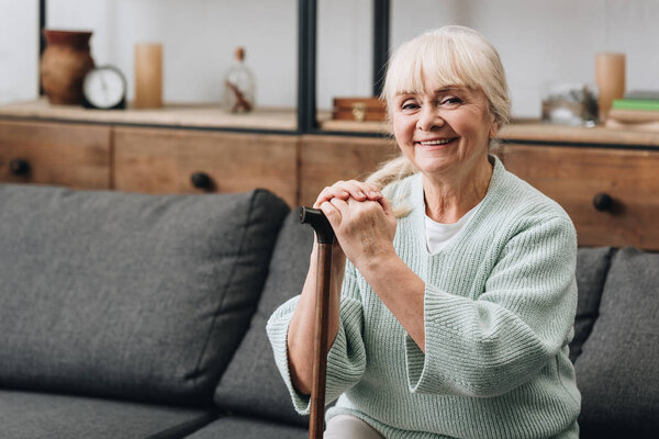cheerful senior woman sitting on sofa and holding walking stick 