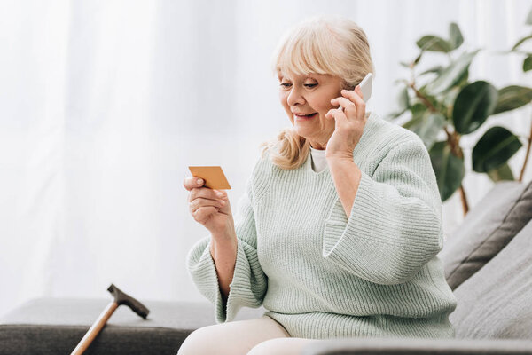 smiling blonde retired woman looking at credit card while talking on smartphone