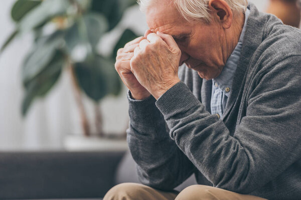 selective focus of upset retired man holding head while sitting on sofa 