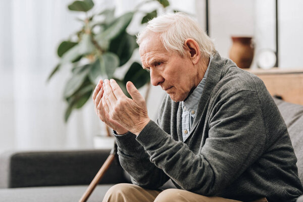  pensioner with grey hair looking at hands while sitting on sofa 