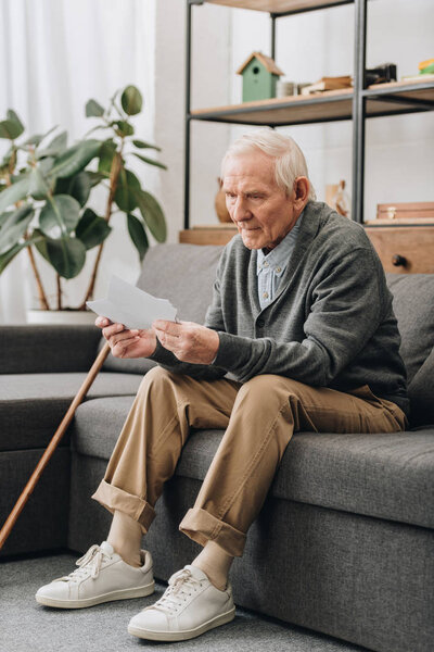sad pensioner with grey hair looking at photos while sitting on sofa 