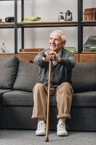 cheerful pensioner smiling and holding walking stick and sitting on sofa