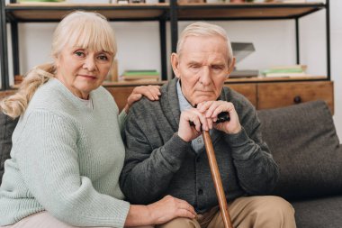 supportive senior wife sitting near retired husband with walking cane in living room  clipart