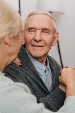 selective focus of smiling pensioner looking at senior wife at home  clipart