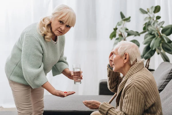 senior woman giving pills and glass of water to old man and looking at camera