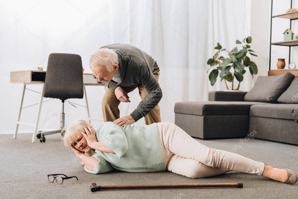 supportive retired husband looking at senior wife lying on floor and holding head 