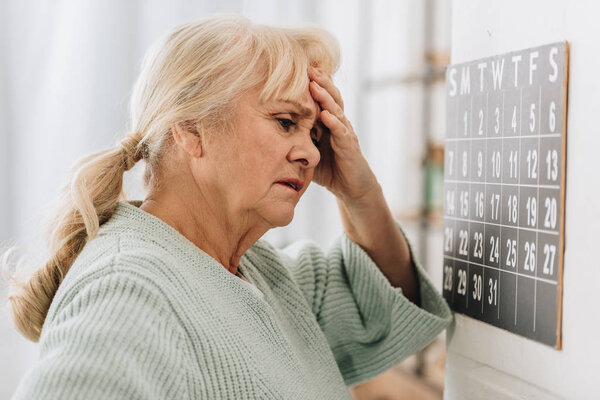 upset woman with gray hair touching head and looking at wall calendar