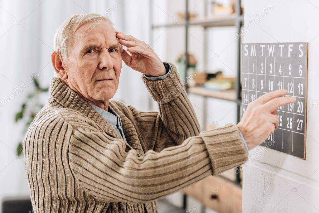 senior man touching wall calendar and head while looking at camera