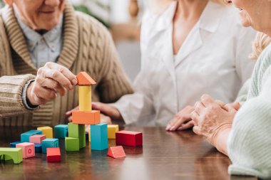 cropped view of caregiver sitting with retired man and woman and playing with wooden toys  clipart