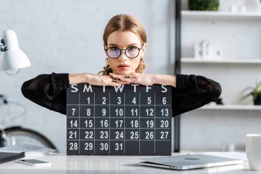 beautiful businesswoman in black clothes and glasses sitting on chair and holding a calendar clipart