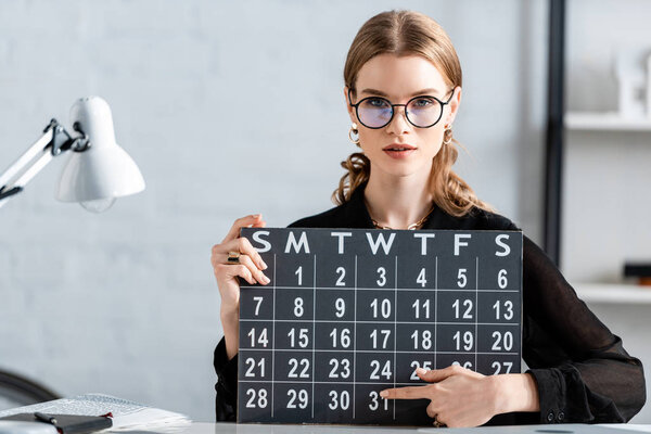 businesswoman in black clothes and glasses holding calendar and sitting on chair
