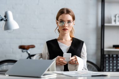 işkadını formal giyim ve bilgisayar masa başında oturan ve işyerinde smartphone kullanarak gözlük