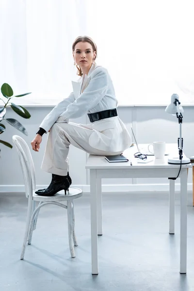 フォーマルウェアの机の上に座っていると 職場でカメラ目線のスタイリッシュな女性実業家 — ストック写真