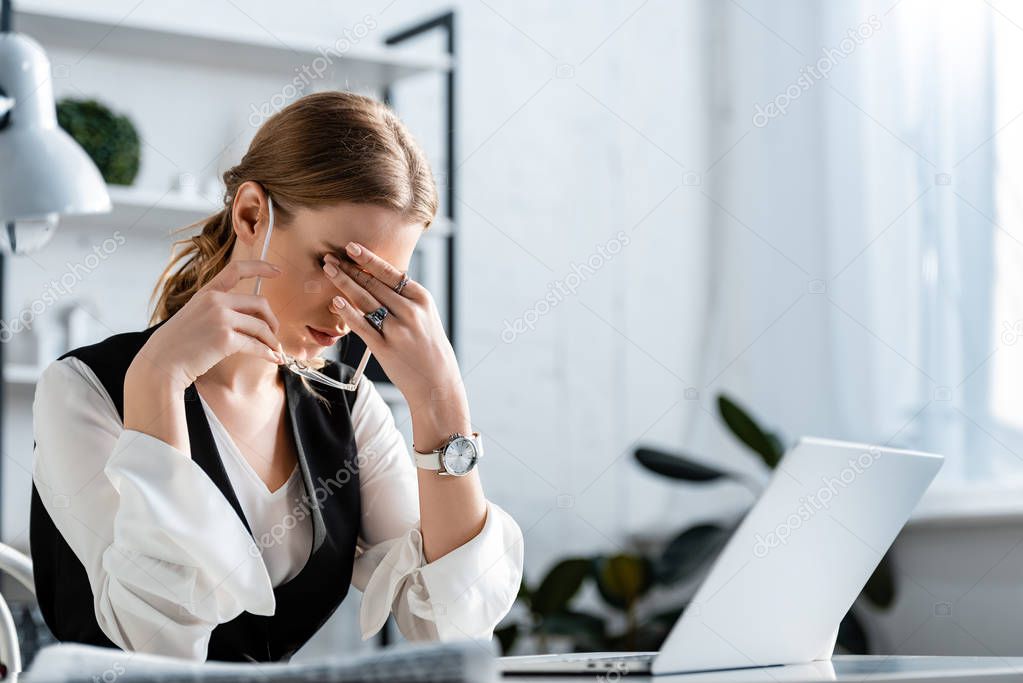 businesswoman in formal wear sitting at computer desk, touching forehead and suffering from headache at workplace