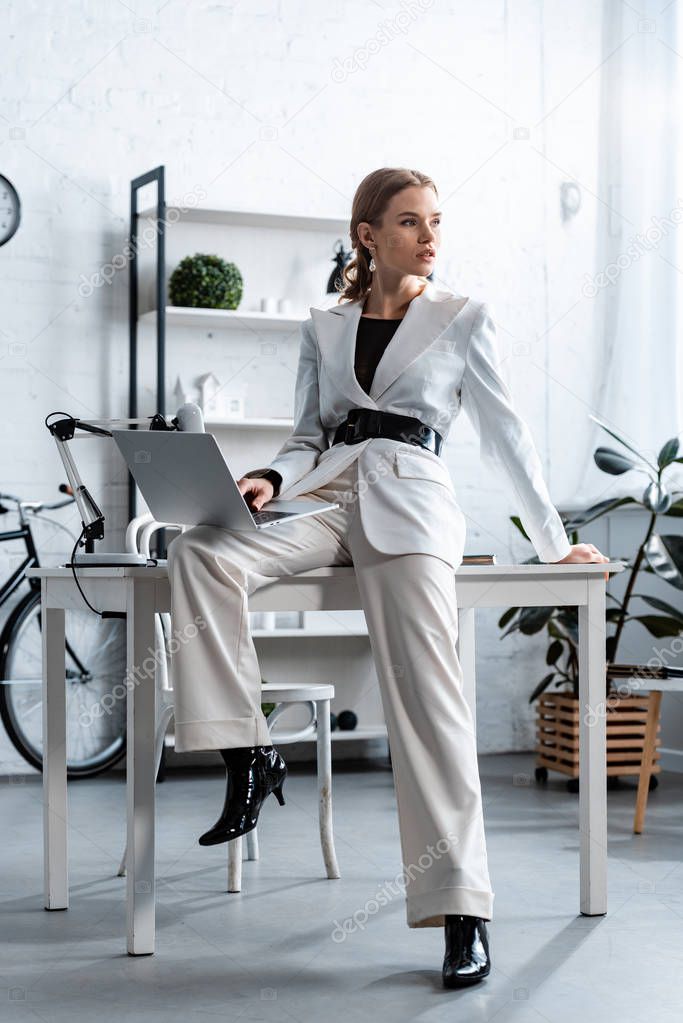businesswoman in white formal wear sitting on desk with laptop in office and looking away