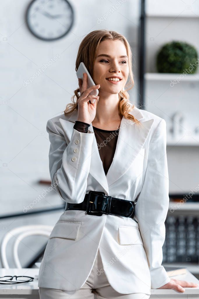 beautiful smiling businesswoman in white formal wear talking on smartphone at workplace