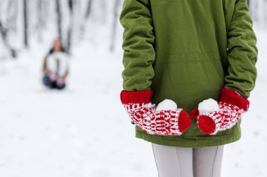 cropped view of child holding snowballs behind back and mother at background in winter park clipart