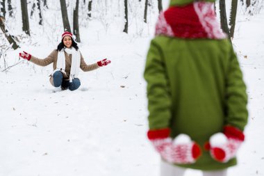 cropped view of child holding snowballs behind back and smiling african american mother at background in winter park clipart
