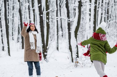 cheerful african american mom playing snowballs with daughter in winter park clipart