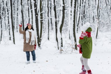 cheerful african american mom smiling and playing snowballs with daughter in warm clothing in winter park clipart