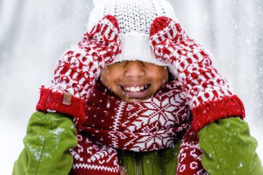cute african american child with knitted hat pulled over eyes smiling during snowfall clipart