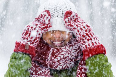 close up view of smiling african american child with knitted hat pulled over eyes during snowfall clipart