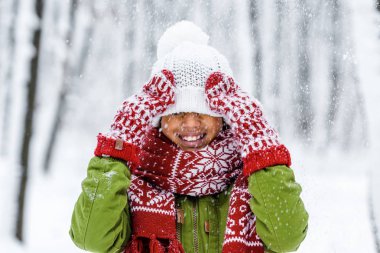 smiling african american child with knitted hat pulled over eyes during snowfall in winter park clipart