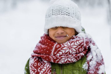 smiling african american child in warm clothing with knitted hat pulled over eyes in winter clipart