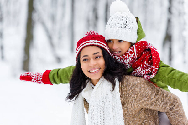 beautiful african american woman giving piggyback ride to cheerful daughter in winter park