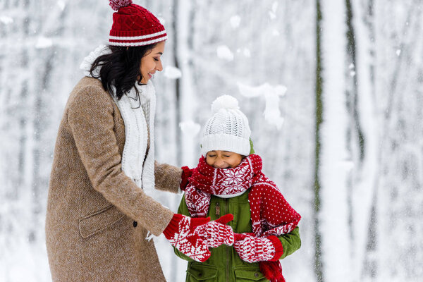 cheerful african american mom hugging cute child with closed eyes during snowfall in winter park