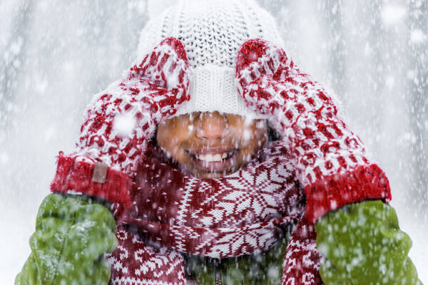 close up view of smiling african american child with knitted hat pulled over eyes during snowfall